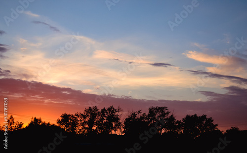 Morning glow illuminates the clouds on a beautiful colorful sky above the silhouettes of trees at dawn