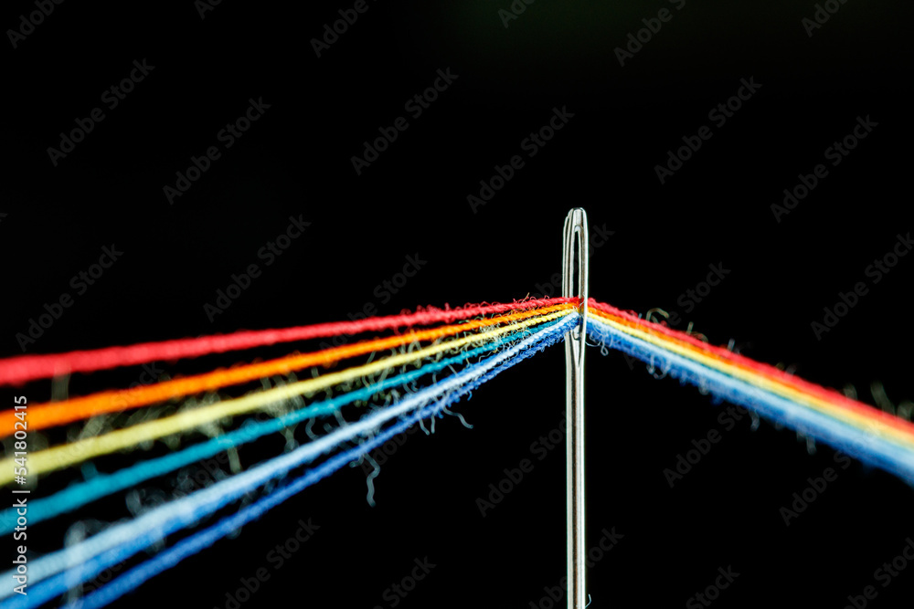 Obraz premium multi-colored threads for sewing in the form of a rainbow pass through an antique needle on a black background
