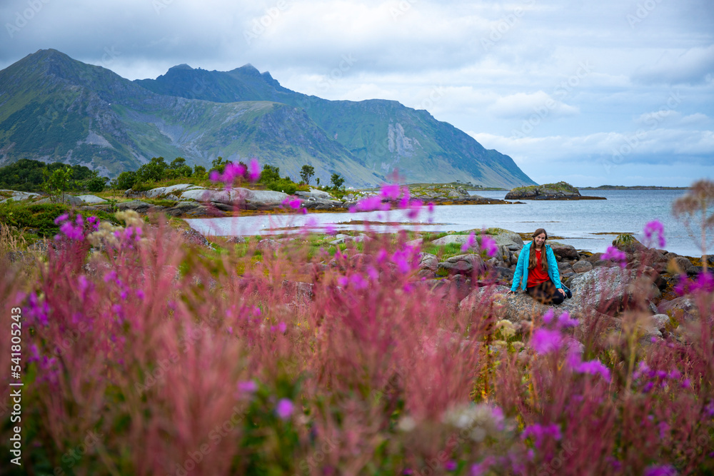 A beautiful girl sits on moss surrounded by lush purple flowers with huge mountains in the background; lofoten islands, norway and its fjords