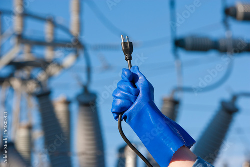 Fototapeta A gloved hand holds wires and plugs in front of an electrical utility substation