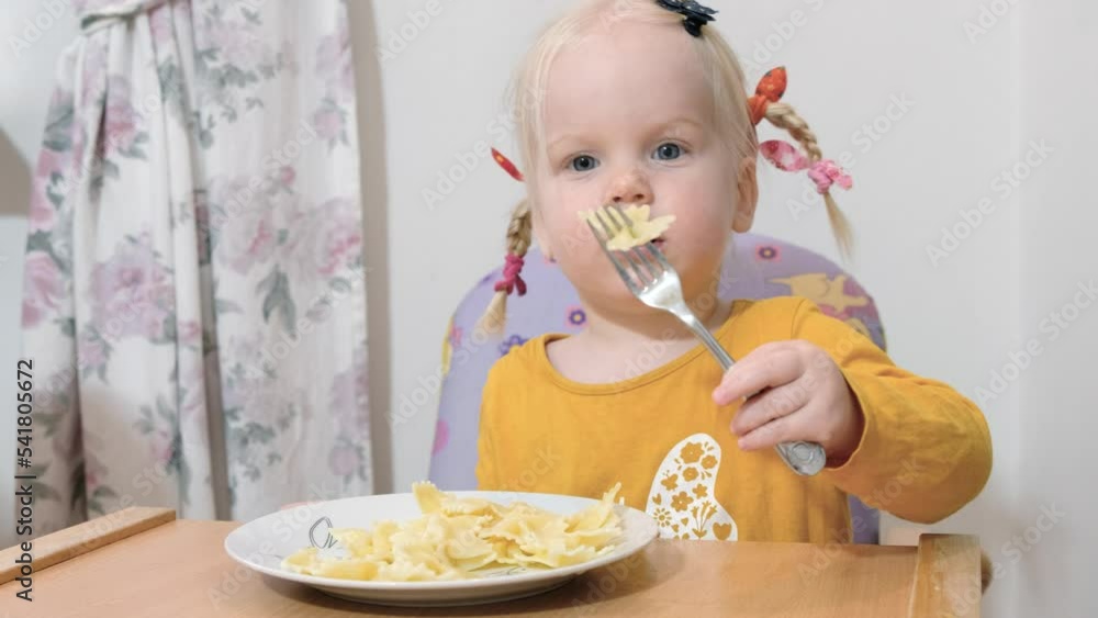 A girl with pigtails is sitting at the table and eating Farfalle pasta with a fork. A girl with pigtails is sitting at the table and eating Farfalle pasta with a fork.