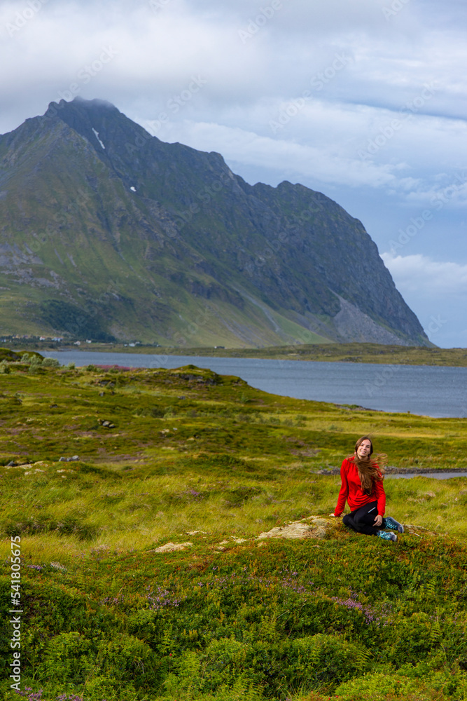 girl in red sweatshirt sits on moss under huge cliffs in the Norwegian fjords, panorama of lofoten islands landscape, norway,