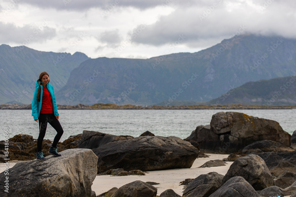 girl in blue jacket walks on the beach with huge rocks and mountains in the background, the Lofoten islands in norway and their harsh landscapes