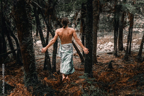 caucasian young man with bare back and legs covered with blanket jumping with arms outstretched between forest wonders on waitawheta tramway, new zealand