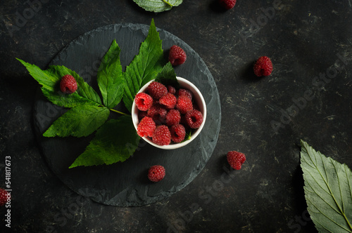 Ripe fresh raspberries and raspberry leaves on a black slate stand, against a black background. Top view, natural lighting, in selective focus with copy space
