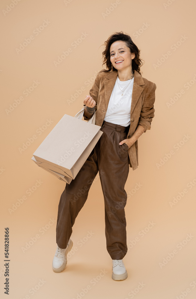 full length of cheerful woman with shopping bags posing in suede jacket and brown pants on beige background full length of cheerful woman with shopping bags posing in suede jacket and brown pants on beige background