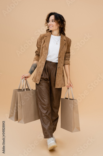 joyful woman in suede jacket walking with shopping bags and looking away on beige background
