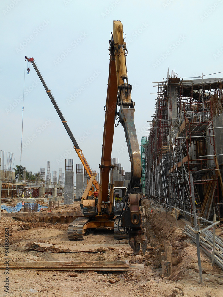 SELANGOR, MALAYSIA –MARCH 15, 2016: Sheet pile cofferdam driven machine at the construction site. The machine drove the sheet pile to the earth using vibrated hydraulic arm. Workers control. SELANGOR, MALAYSIA –MARCH 15, 2016: Sheet pile cofferdam driven machine at the construction site. The machine drove the sheet pile to the earth using vibrated hydraulic arm. Workers control.