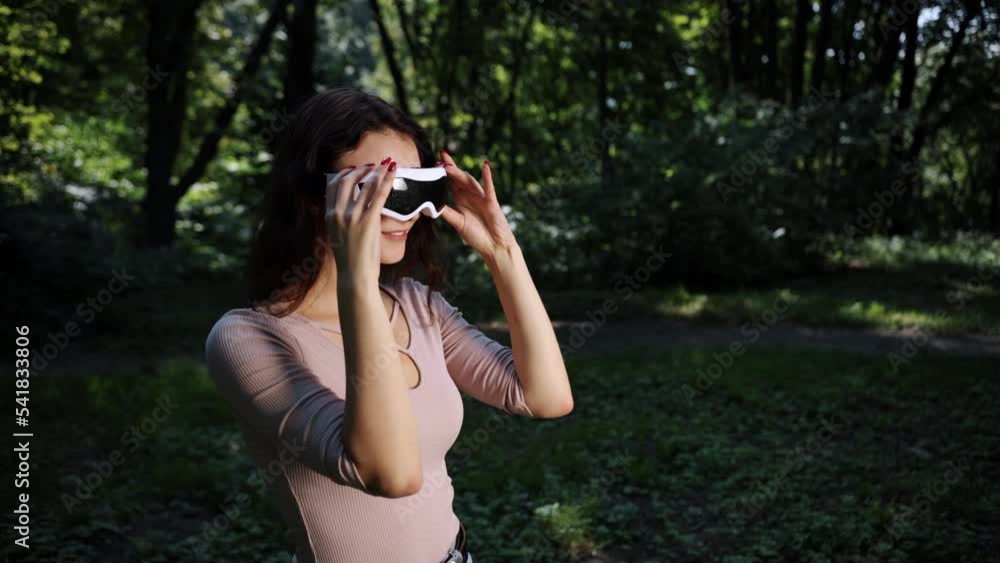 Background of blurry nature with people and technology, young woman playing games in the park while wearing VR glasses at sunset against the blurred background of nature Background of blurry nature with people and technology, young woman playing games in the park while wearing VR glasses at sunset against the blurred background of nature