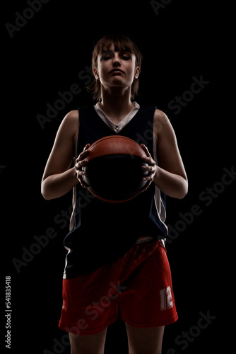 Female basketball player. Beautiful girl holding ball. Side lit half silhouette studio portrait against black background. © Nikola Spasenoski