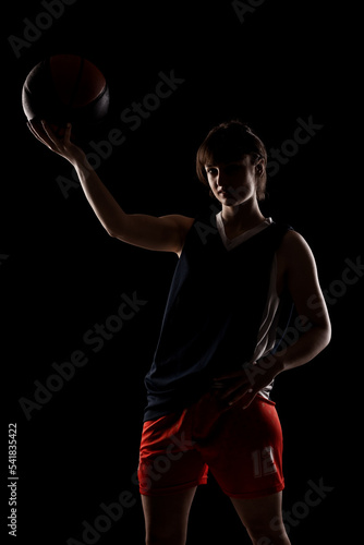 Female basketball player. Beautiful girl holding ball. Side lit half silhouette studio portrait against black background. © Nikola Spasenoski