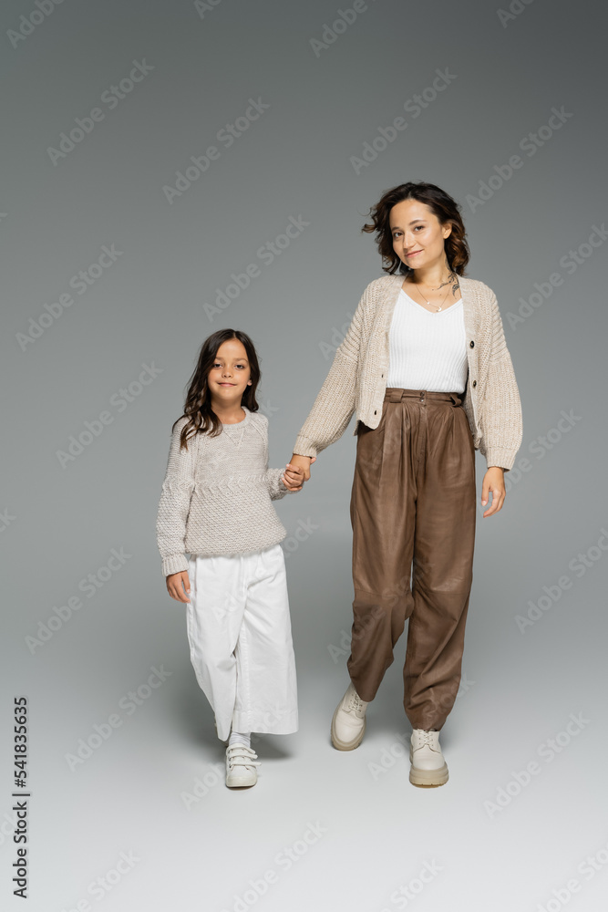 mother with child in stylish autumn outfit holding hands and looking at camera while walking on grey background mother with child in stylish autumn outfit holding hands and looking at camera while walking on grey background