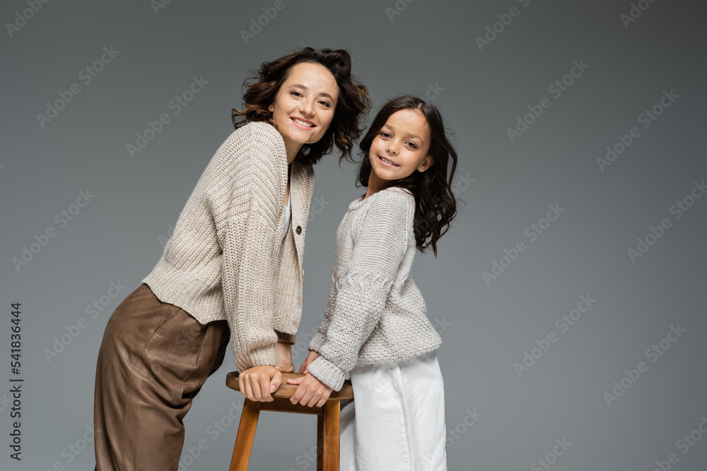 smiling mother and daughter in trendy autumn outfit posing near wooden stool isolated on grey smiling mother and daughter in trendy autumn outfit posing near wooden stool isolated on grey