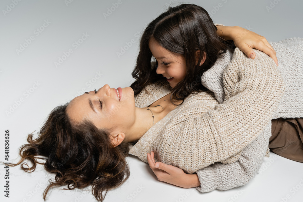 smiling woman in warm cardigan embracing daughter while lying on grey background smiling woman in warm cardigan embracing daughter while lying on grey background