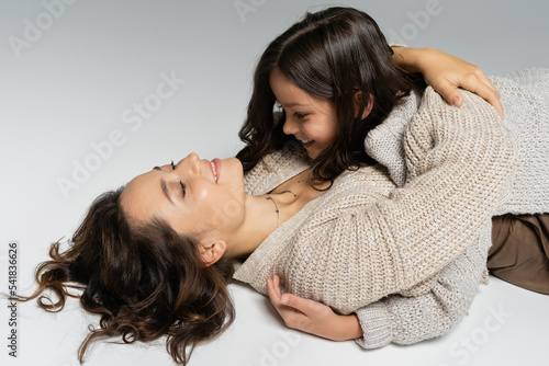 smiling woman in warm cardigan embracing daughter while lying on grey background