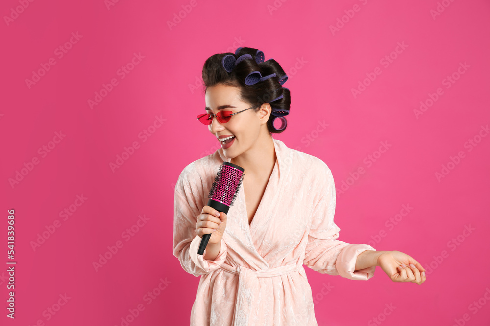 Happy young woman in bathrobe with hair curlers and sunglasses singing into hairbrush on pink background