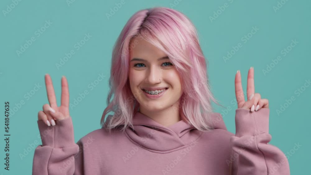 Excited young woman with pink hair color doing peace symbol with fingers, smiling at camera. Positive student showing optimistic gesture, having friendly expression. Person expressing peaceful V sign Excited young woman with pink hair color doing peace symbol with fingers, smiling at camera. Positive student showing optimistic gesture, having friendly expression. Person expressing peaceful V sign