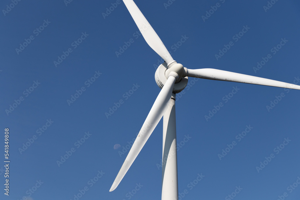 A large wind turbine is seen on a clear, blue sky day with the moon faintly seen in the background. A large wind turbine is seen on a clear, blue sky day with the moon faintly seen in the background.