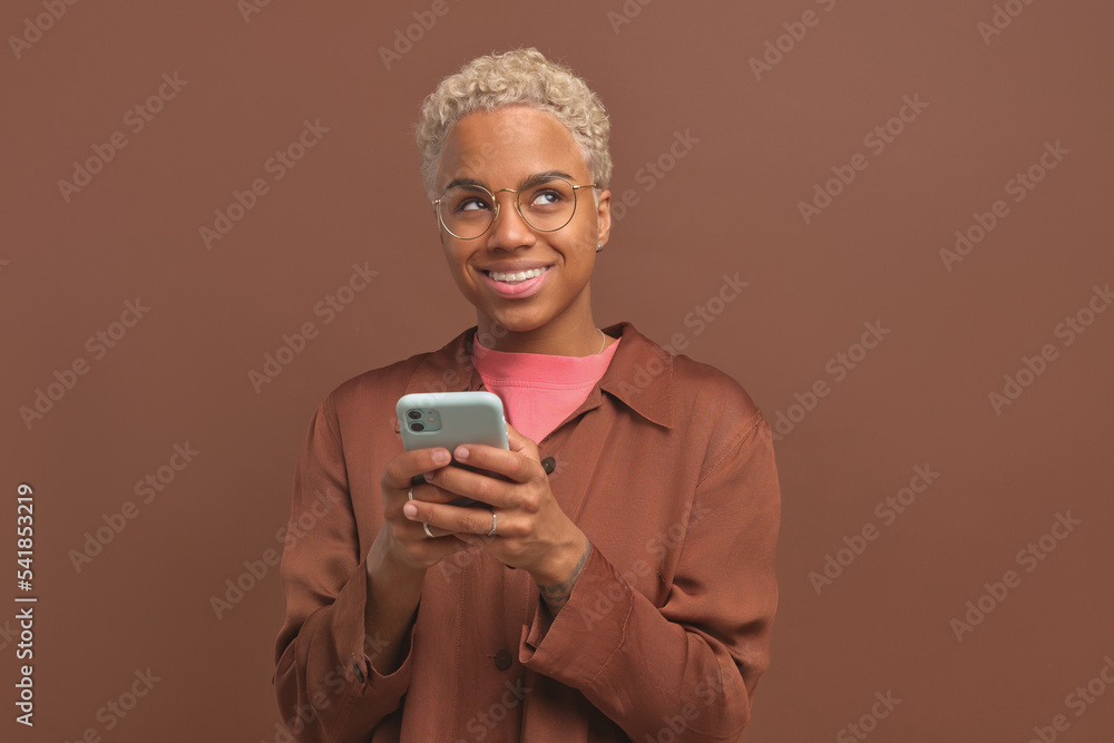 Fototapeta premium Young shy pretty African American woman in glasses with mobile phone in hands dreamily looks up and chats on Internet with new friend from social network stands in brown studio. Smartphone, apps