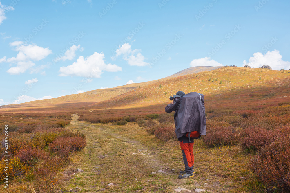 Alone traveler in raincoat with large backpack photographs nature among fading autumn vegetations on high mountain plateau. Backpacker with photo camera shoots mountain landscape in changeable weather