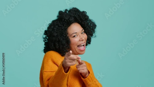 Hey you finger gesture, Profile portrait of African American woman smiling at camera and indicating happily at camera Gotcha expression, choosing and smiling, blue studio background, slow motion copy
