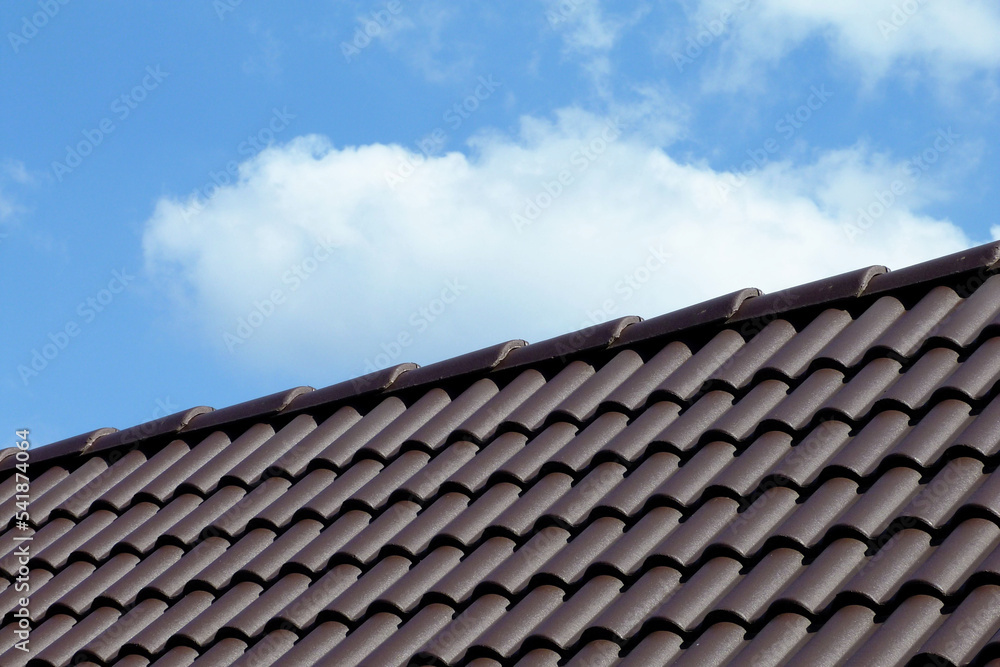 sloped dark brown clay tile roof and ridge. construction, modern building materials and technology concept. blue sky and fluffy white cloud. bright summer light. low angle abstract view 