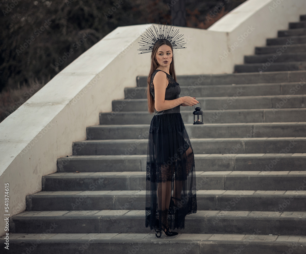 Girl in a black crown and black dress. Stands on the steps of the stairs with a lantern in his hands. Girl in a black crown and black dress. Stands on the steps of the stairs with a lantern in his hands.