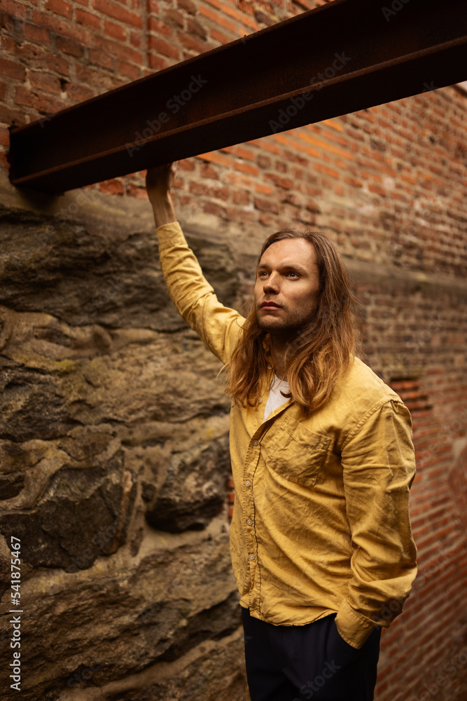 A caucasian man with long hair in a yellow shirt and blue pants standing infront of a brick wall