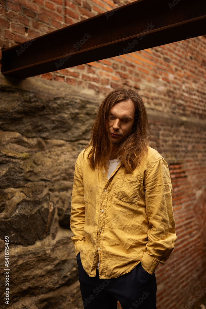A sad caucasian man with long hair in a yellow shirt and blue pants standing infront of a brick wall