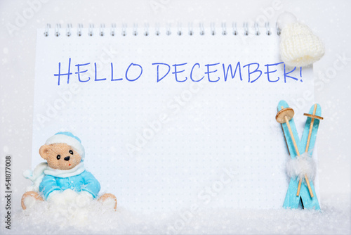 HELLO DECEMBER. Teddy bear in blue clothes sitting on a sled, blue wooden skis, a white hat and a notebook with the words HELLO DECEMBER on a background of white snow. photo