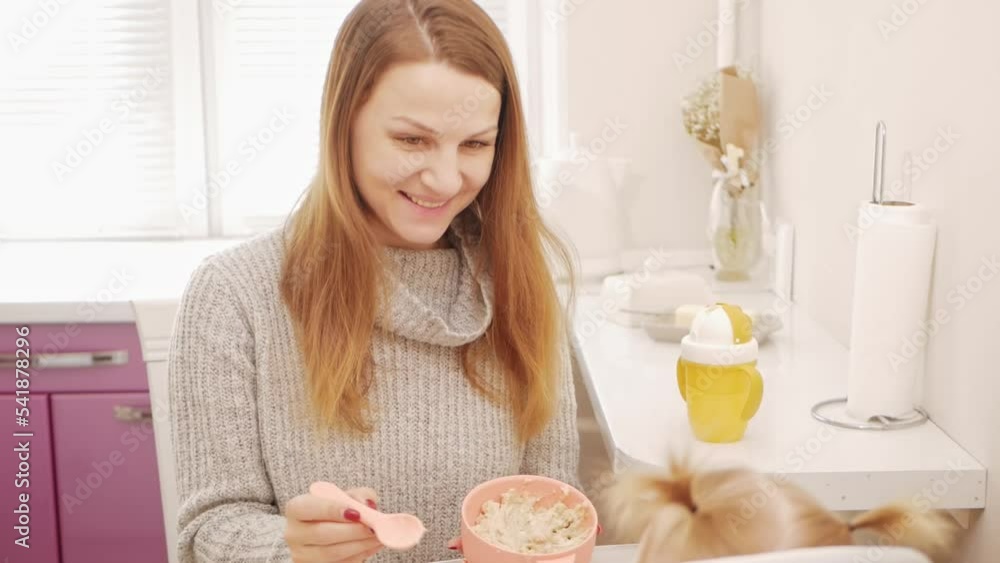 mother feeds her daughter porridge, one-year-old child eats with spoon in kitchen, love and care, funny. brunette mother feeds her daughter porridge, one-year-old child eats with spoon in kitchen, love and care, funny. brunette