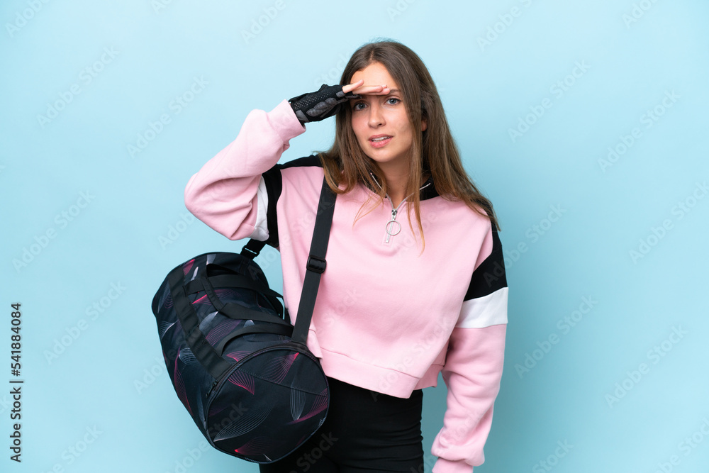Young sport woman with sport bag isolated on blue background looking far away with hand to look something Young sport woman with sport bag isolated on blue background looking far away with hand to look something