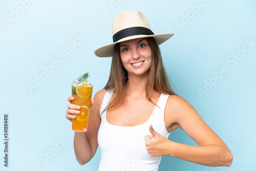 Young beautiful woman holding a cocktail isolated on blue background with thumbs up because something good has happened
