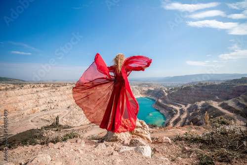 Side view of a beautiful sensual woman in a red long dress posing on a rock high above the lake in the afternoon. Against the background of the blue sky and the lake in the form of a heart