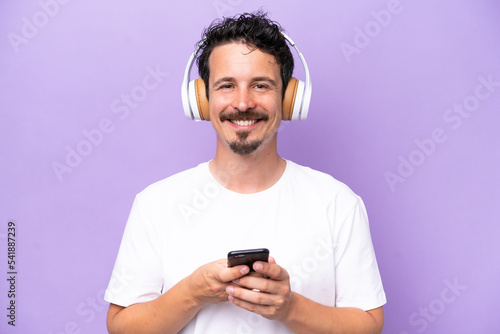 Young caucasian man isolated on purple background listening music with a mobile and looking front