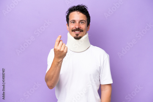 Young caucasian man wearing neck brace isolated on purple background doing coming gesture