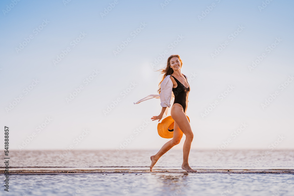 Naklejka premium Running young gorgeous woman in black swimsuit with fluttering hair, white shirt rising hat up spending time on the beach smiles wide. Joyful vibes at summertime vacations. Beautiful Swedish girl
