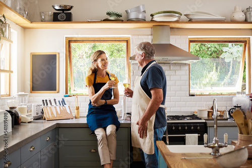 Love, wine and mature couple in kitchen cooking, drinking and spending free time together in home. Retirement, happy man and woman with wine glass, smile and apron relax before dinner in modern house