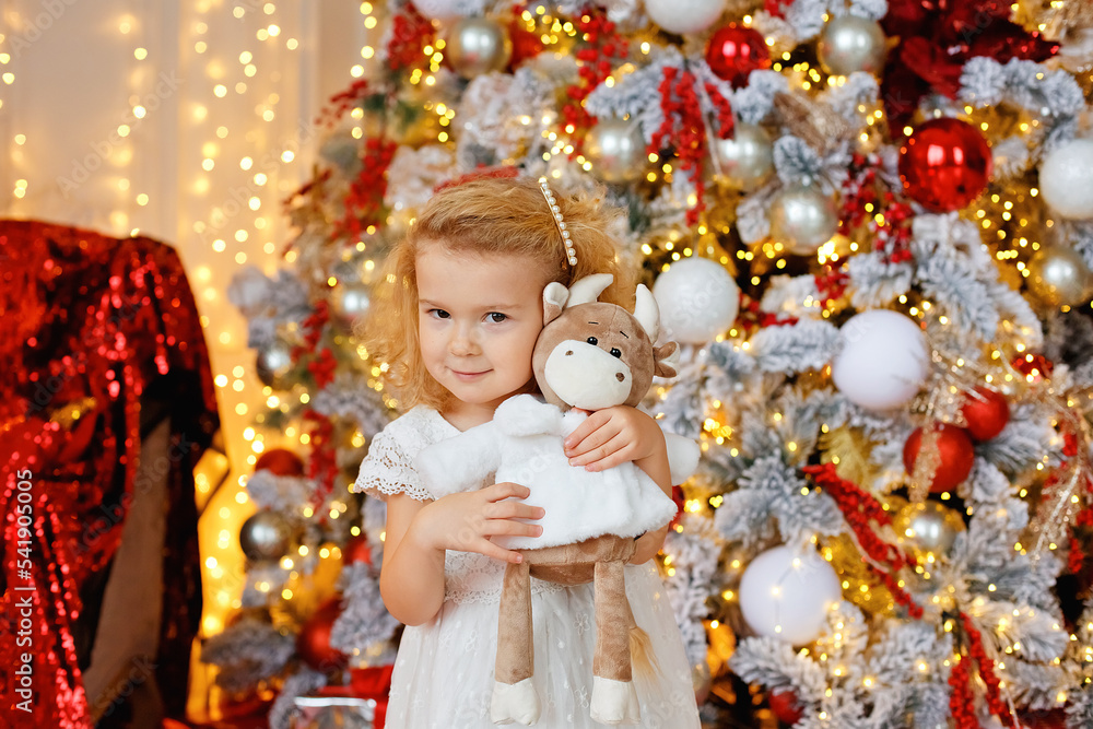 A large portrait of a small curly blonde girl in a white dress and a soft toy in her hands against the background of bokeh lights and a decorated Christmas tree. A large portrait of a small curly blonde girl in a white dress and a soft toy in her hands against the background of bokeh lights and a decorated Christmas tree.