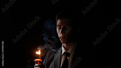 A serious young man lights his pipe with a match, a man in a gray suit with a tie holds a burning match in his hand, deep shadows in a retro noir style photo