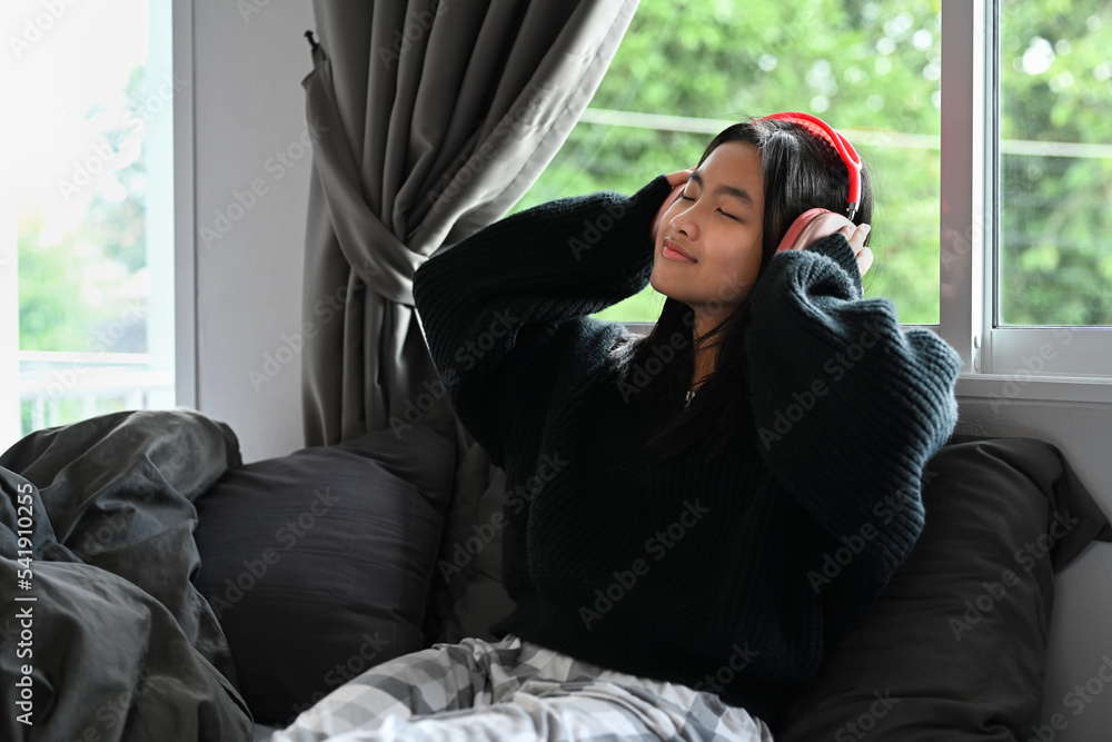 Obraz premium Cheerful young woman listening to music on wireless headphone while sitting in bedroom
