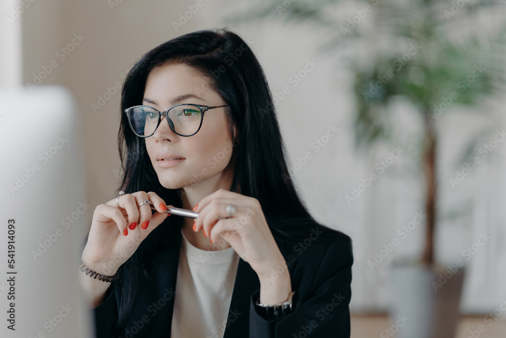 Skilled female freelancer concentrated at computer display, wears optical glasses, holds pen, has dark hair, makes online banking. Business lady works distantly from home, develops new startup project Skilled female freelancer concentrated at computer display, wears optical glasses, holds pen, has dark hair, makes online banking. Business lady works distantly from home, develops new startup project