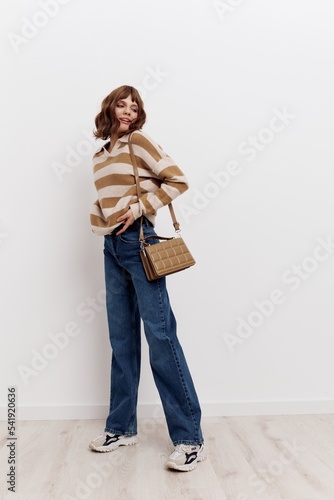 a full-length photo of stylish woman standing on a light background in a striped sweater and blue jeans, posing relaxed with a beige bag worn over her shoulder, adjusting the sweater and looking away