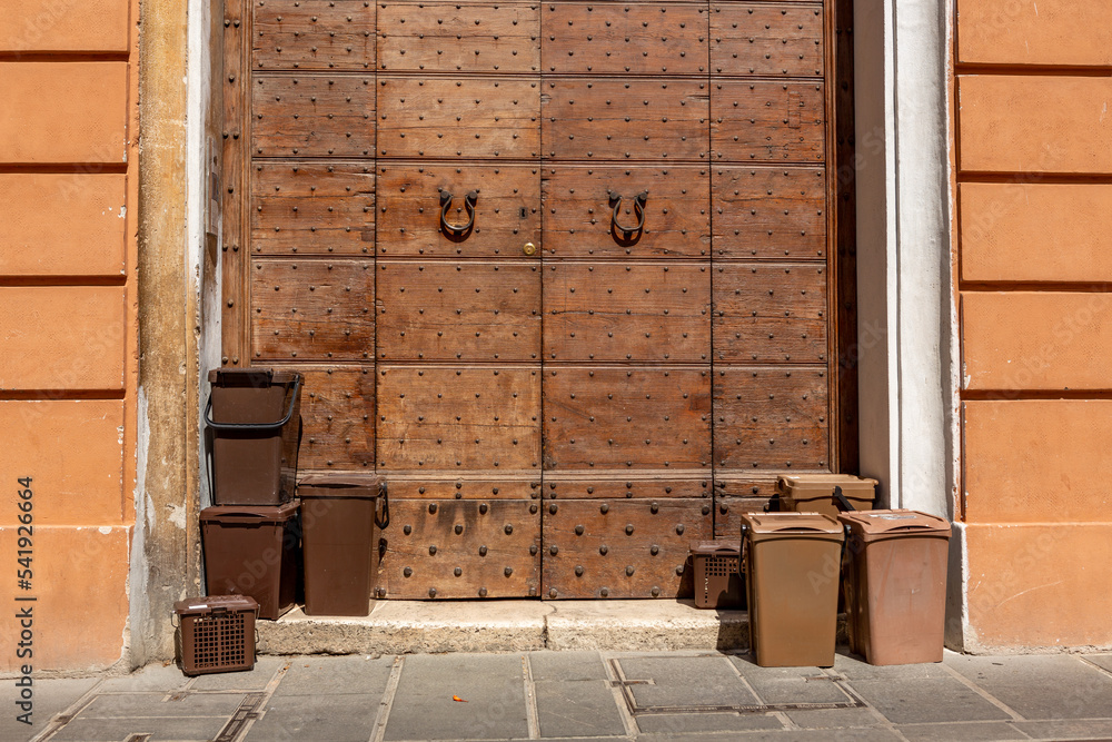 Obraz premium Brown plastic bins with biological waste on the sidewalk in front of a historic old wooden door in the village of Foligno in the central Italian region of Umbria in the province of Perugia.