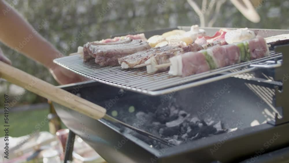 Barbecue grill with meat and man stirring coal with fork and blowing out fire. Close-up of vegetables, mushroom and shashlik being roasted outdoors. Barbecue party concept Barbecue grill with meat and man stirring coal with fork and blowing out fire. Close-up of vegetables, mushroom and shashlik being roasted outdoors. Barbecue party concept