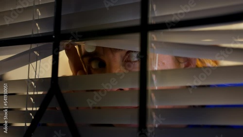 A paranoid scared woman looking through blinds at home. The girl is anxious and suspicious as she is looking out from her home into the street at night