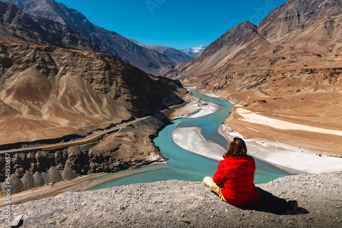 Man in red jacket sit on cliff and look at Sangam or confluence of Indus and Zanskar Rivers at Leh, Ladakh, India. photo