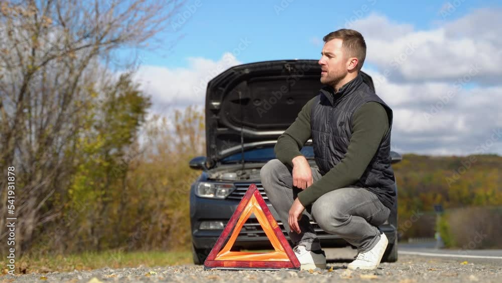 Frustrated man on road with broken car. Side warning triangle, warning oncoming traffic of a broken down car Frustrated man on road with broken car. Side warning triangle, warning oncoming traffic of a broken down car