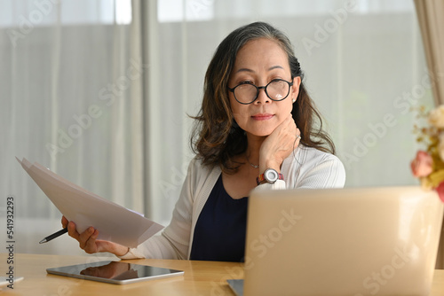 Old woman wearing glasses working on laptop online, sitting at desk on living room.