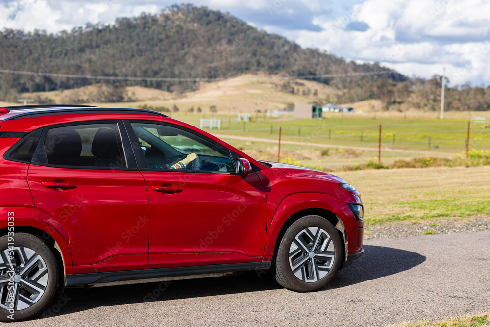 Red electric vehicle car driving down rural country road on sunlit day Red electric vehicle car driving down rural country road on sunlit day